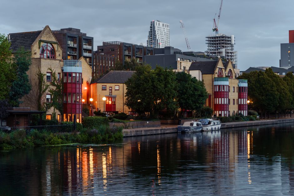 A view of residential buildings along the water's edge in Bromley-by-Bow, with a canal reflecting the illuminated windows and streetlights. The buildings include a mix of traditional brick houses and 