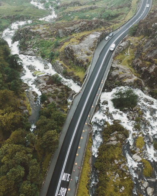 An aerial view of a narrow, winding road with a smooth asphalt surface, bordered by a metal guardrail on one side and a steep rocky hillside on the other, with several vehicles traveling along it. The