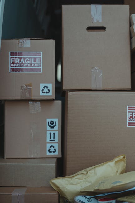 Stacked cardboard boxes of varying sizes inside a property, with some marked as fragile and handling with care, wrapped with clear packing tape, and placed on a wooden surface near a doorway. The boxe