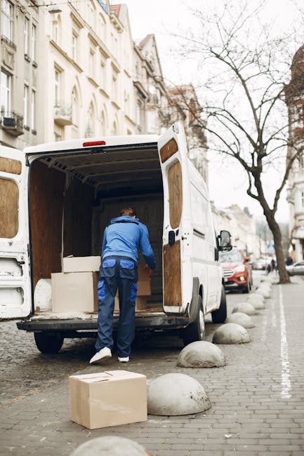 Stacked cardboard boxes of varying sizes inside a property, with some marked as fragile and handling with care, wrapped with clear packing tape, and placed on a wooden surface near a doorway. The boxe